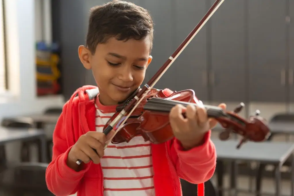 Smiling schoolboy with eyes closed playing violin in classroom at elementary school