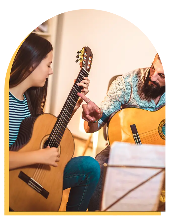 A man teaches a young girl to play the guitar, pointing at the frets as she follows his instructions. Both are holding acoustic guitars, with sheet music in the foreground—capturing a moment from music lessons Westchase families love.