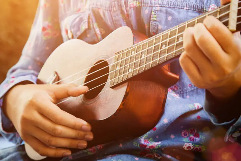 Close-up of a person playing a small wooden ukulele, wearing a blue floral shirt. Warm sunlight shines on their hands and instrument, creating a cozy and relaxed atmosphere.