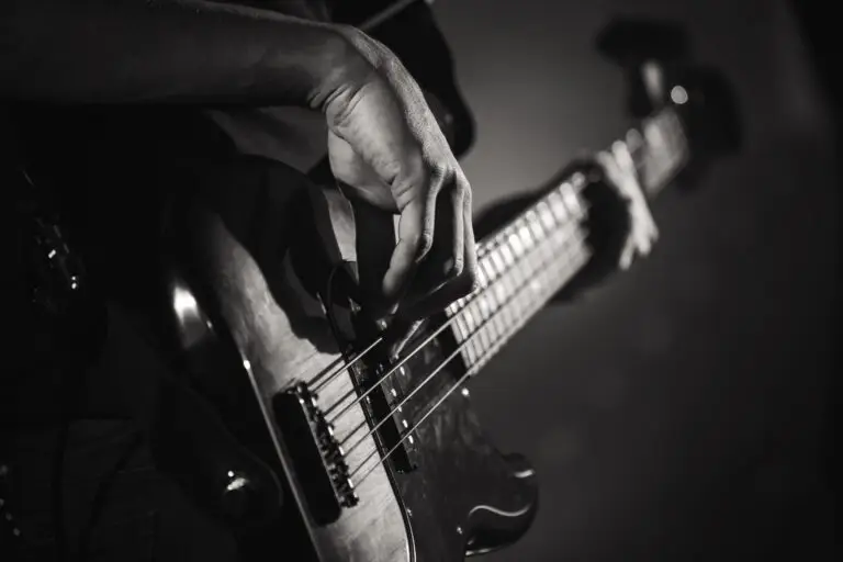 Close-up, black and white photo of a person’s hand playing an electric bass guitar, fingers plucking the strings, with the instrument’s body and neck in focus.