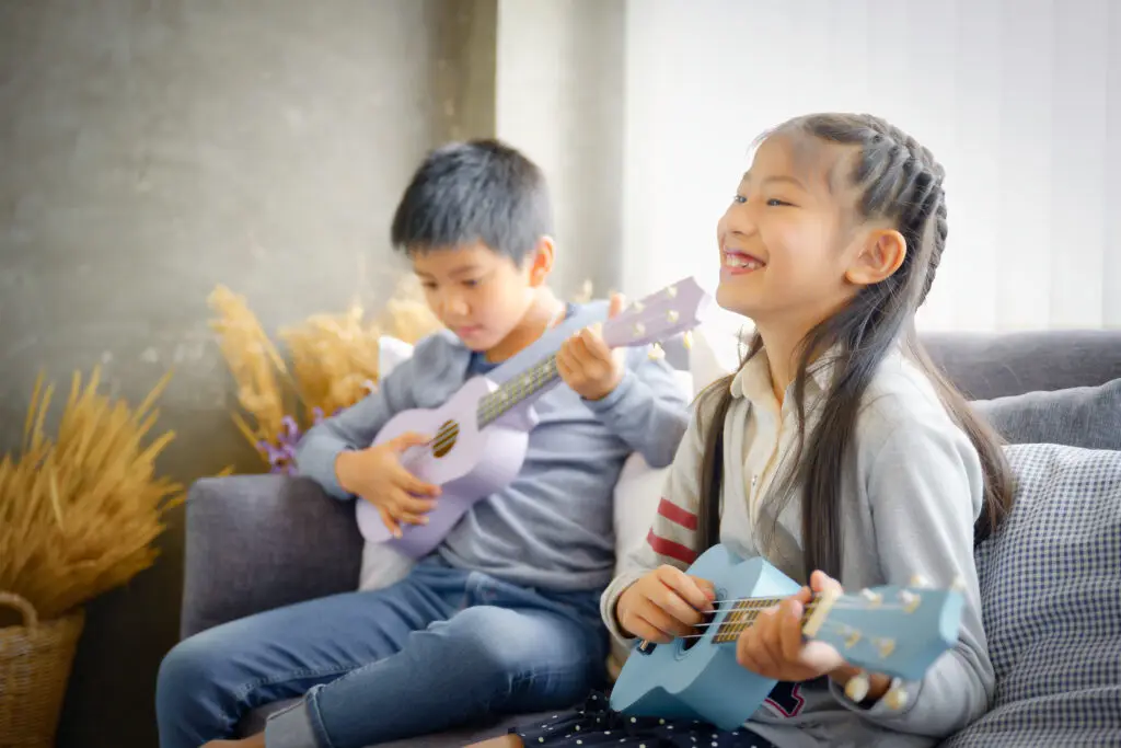 A smiling girl and a boy sit on a couch playing pastel-colored ukuleles; the girl holds a blue one, the boy a purple one. Soft daylight fills the room, and dried plants decorate the background.